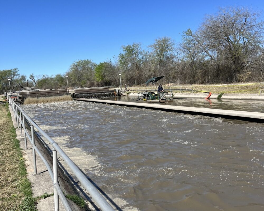 A wastewater treatment facility with a canal undergoing dredging. Machinery and workers under a canopy are seen on one side, while flowing water and a metal railing line the other, surrounded by trees and a bright sky.