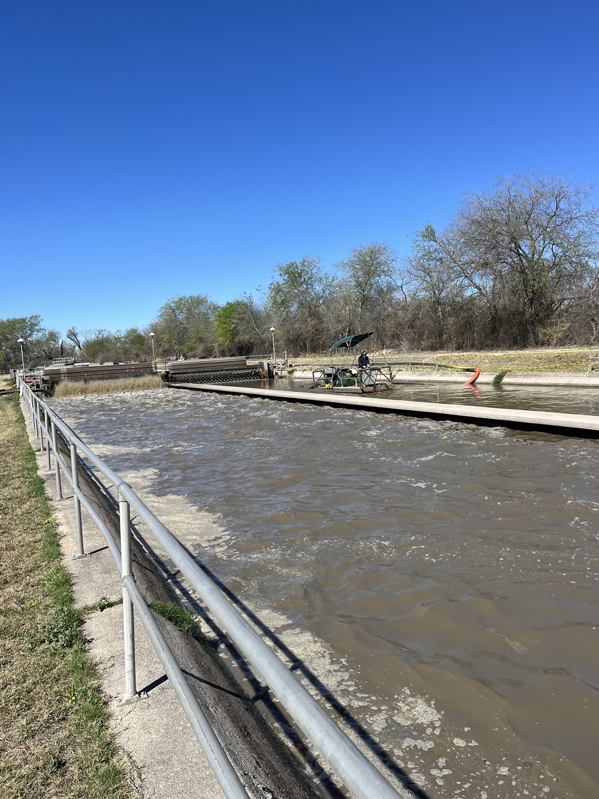 A wastewater treatment facility with a canal undergoing dredging. Machinery and workers under a canopy are seen on one side, while flowing water and a metal railing line the other, surrounded by trees and a bright sky.