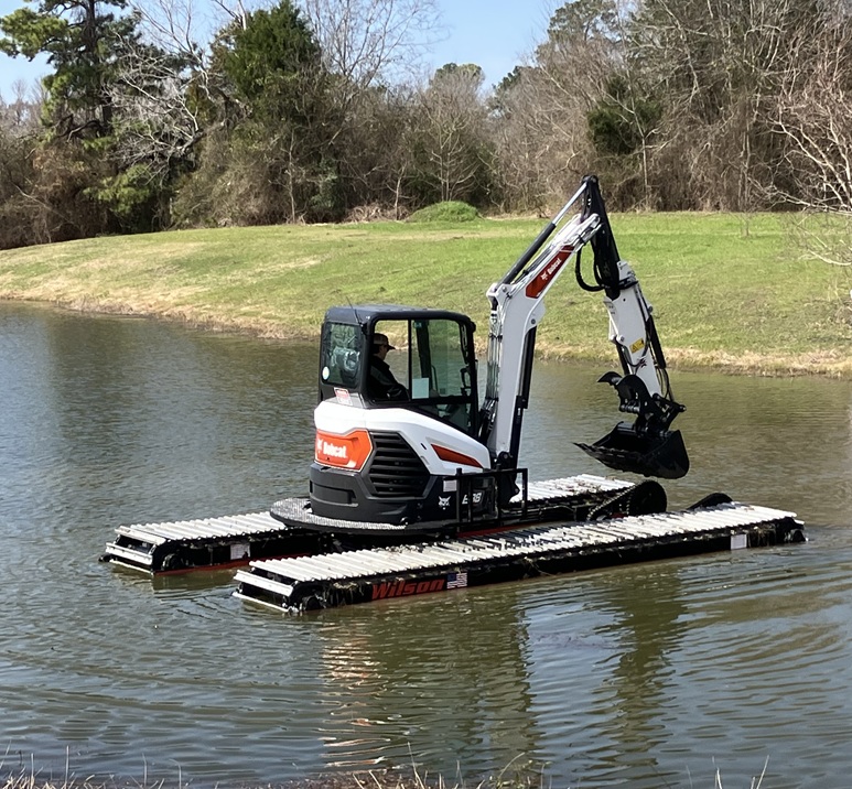 An amphibious excavator equipped with pontoons operating on a pond with its arm extended. The machine demonstrates its adaptability in aquatic environments, surrounded by grassy banks and trees.