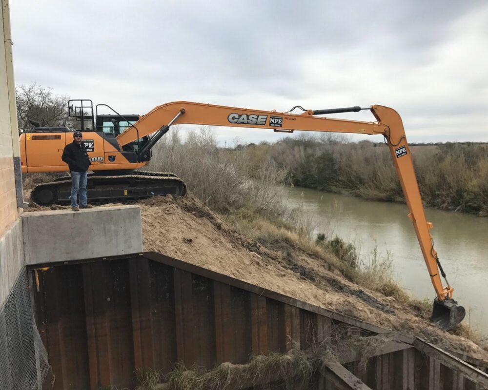 Bullock Construction's large excavator perched on a sandy embankment by a river performing excavation work for a municipality.