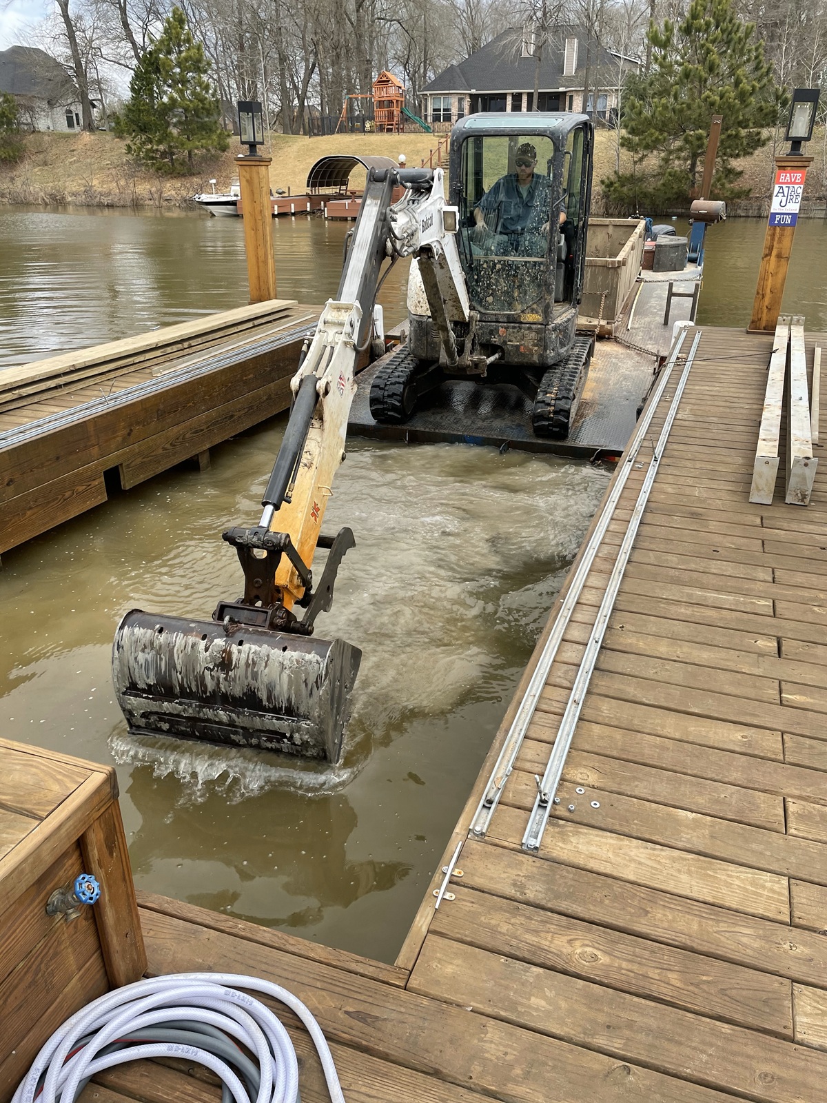 An excavator stationed on a floating platform at a dock, its bucket submerged in the water as part of dredging or maintenance work. Surrounding the platform are wooden docks, trees, and residential houses.