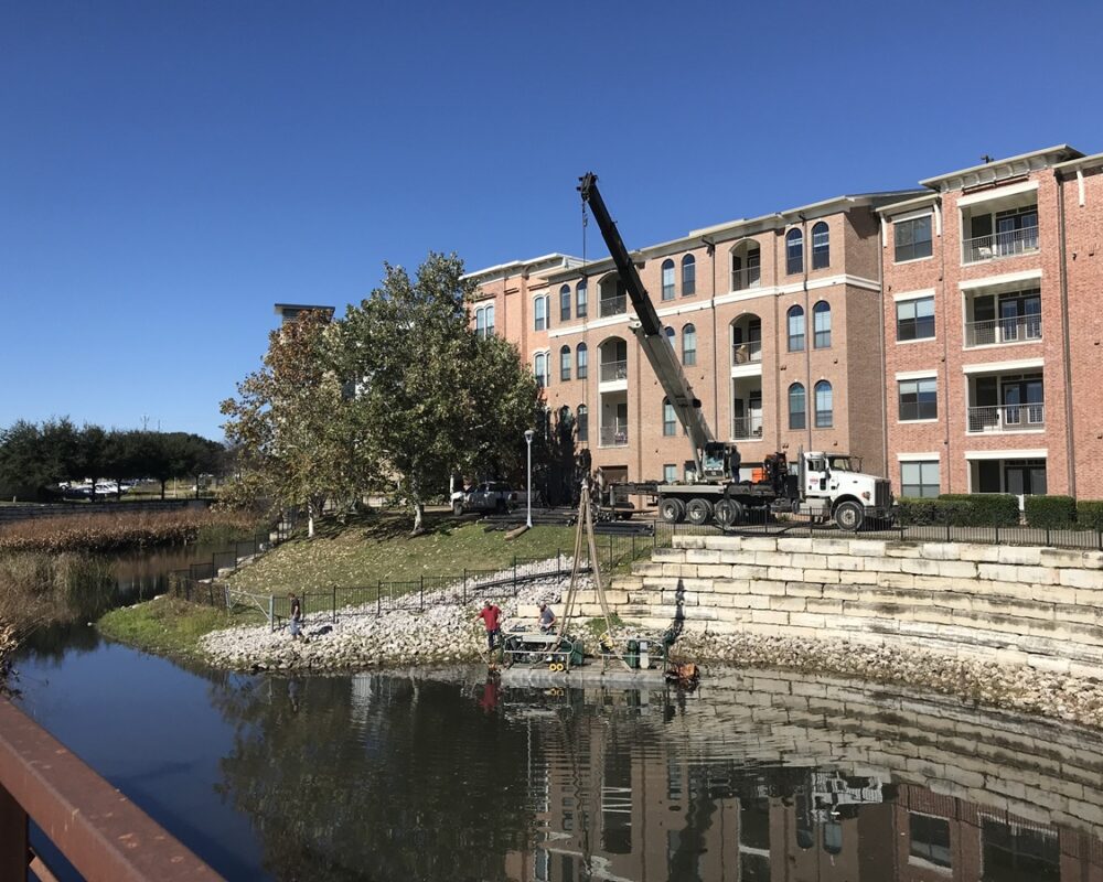 Compact, truck-mounted crane maneuvering along a narrow waterway bank within a residential apartment complex. The machinery is designed for confined spaces, extracting sediment efficiently while minimizing disruption. Apartment buildings with balconies serve as the backdrop, highlighting the urban environment of the dredging operation.