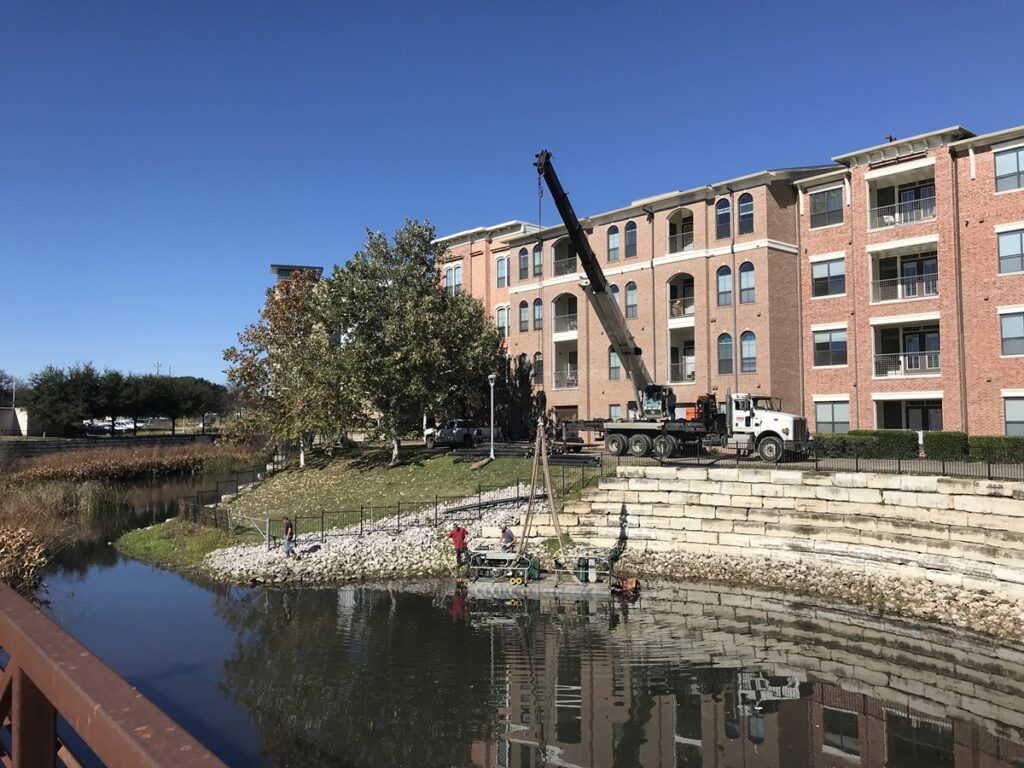 Compact, truck-mounted crane maneuvering along a narrow waterway bank within a residential apartment complex. The machinery is designed for confined spaces, extracting sediment efficiently while minimizing disruption. Apartment buildings with balconies serve as the backdrop, highlighting the urban environment of the dredging operation.