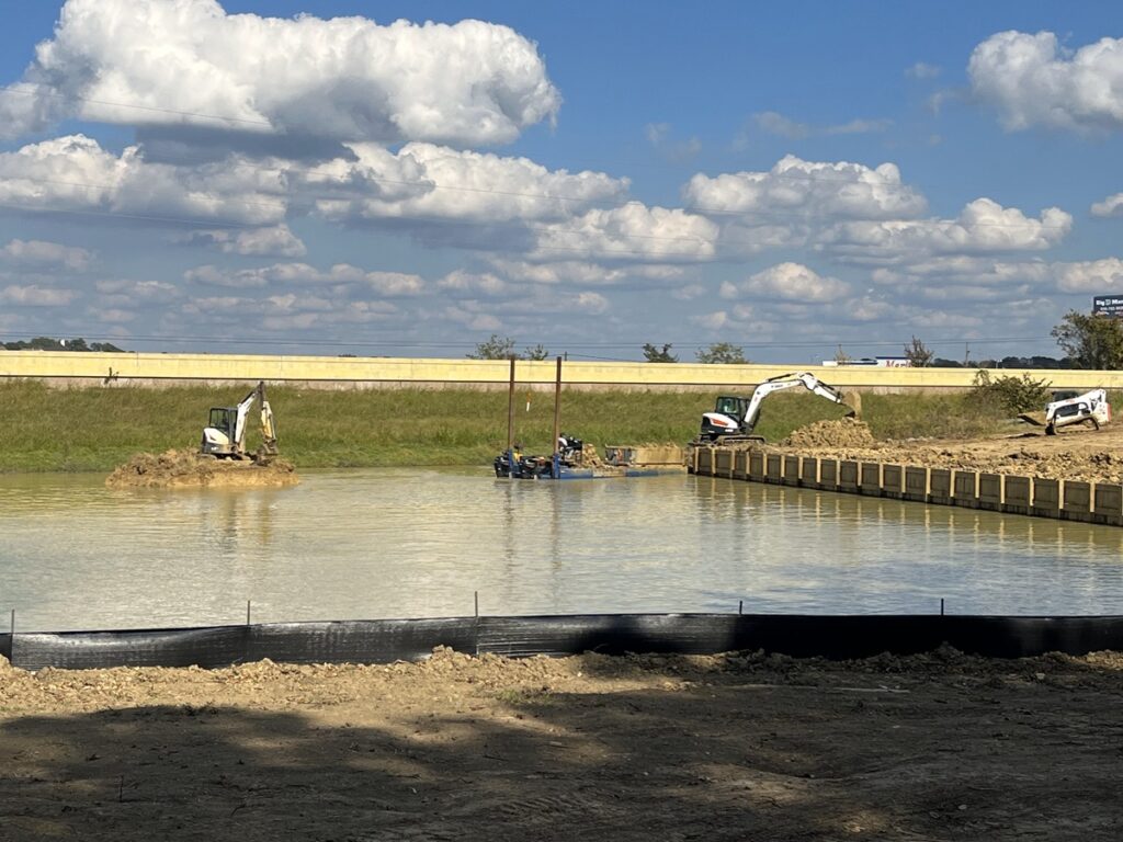 Photo of three excavators involved in dredging operations near a busy highway, including one working from a temporary dirt mound in the water