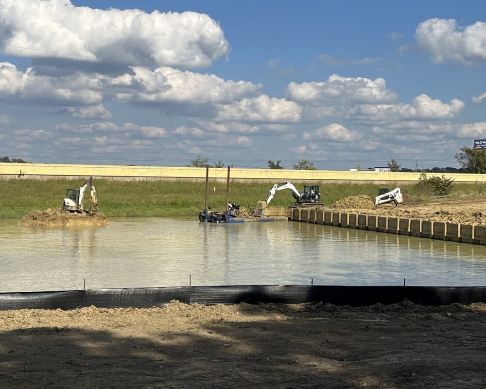 An image of a construction site featuring excavators removing dirt and sediment, with a floating platform and a highway in the background.
