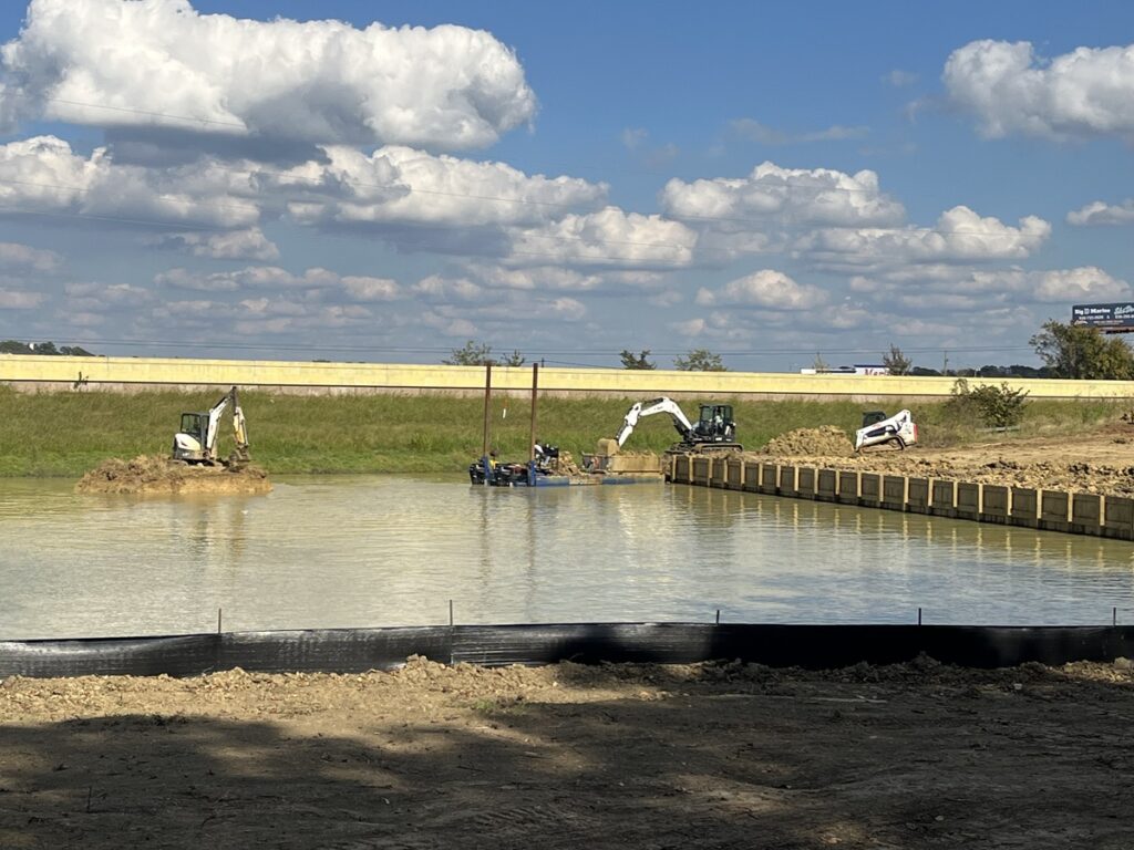 An image of a construction site featuring excavators removing dirt and sediment, with a floating platform and a highway in the background.