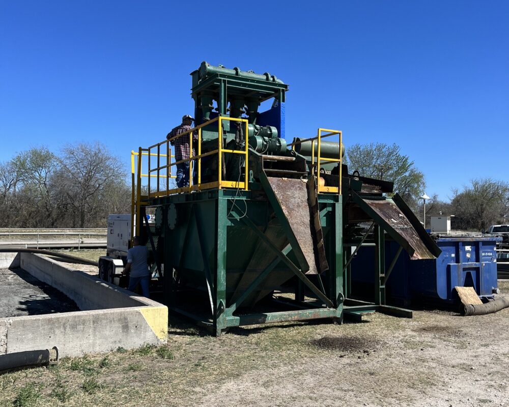 A green industrial dewatering machine with yellow railings, equipped with a conveyor belt system. The machinery is actively separating debris from water at a wastewater treatment facility. Two workers are present, surrounded by dirt, gravel, blue containers, hoses, and trees under a clear blue sky.