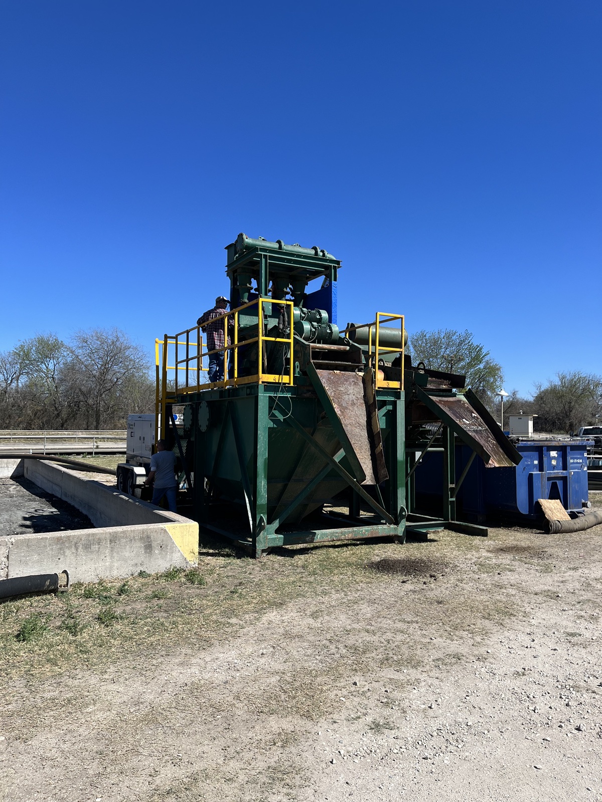 A green industrial dewatering machine with yellow railings, equipped with a conveyor belt system. The machinery is actively separating debris from water at a wastewater treatment facility. Two workers are present, surrounded by dirt, gravel, blue containers, hoses, and trees under a clear blue sky.
