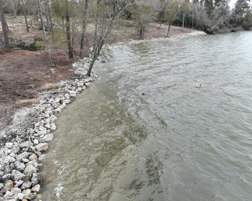 Shoreline of a calm lake with rocks installed as erosion control measures, backed by a forested landscape.