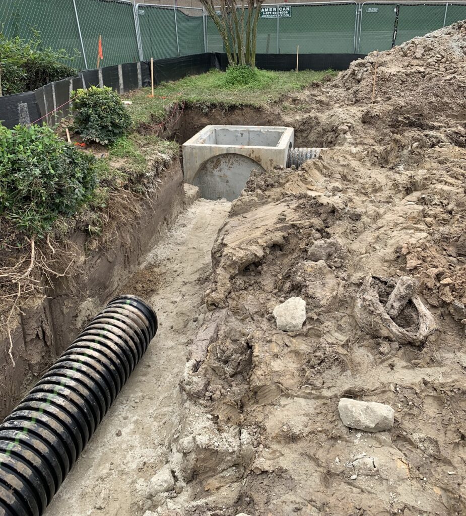 Construction site showing a partially buried concrete structure connected to a corrugated black pipe. The surrounding area is excavated, with dirt and rocks piled around. Green fences and black barriers appear in the background, along with vegetation and construction markers. This highlights drainage or utility installation at the site."