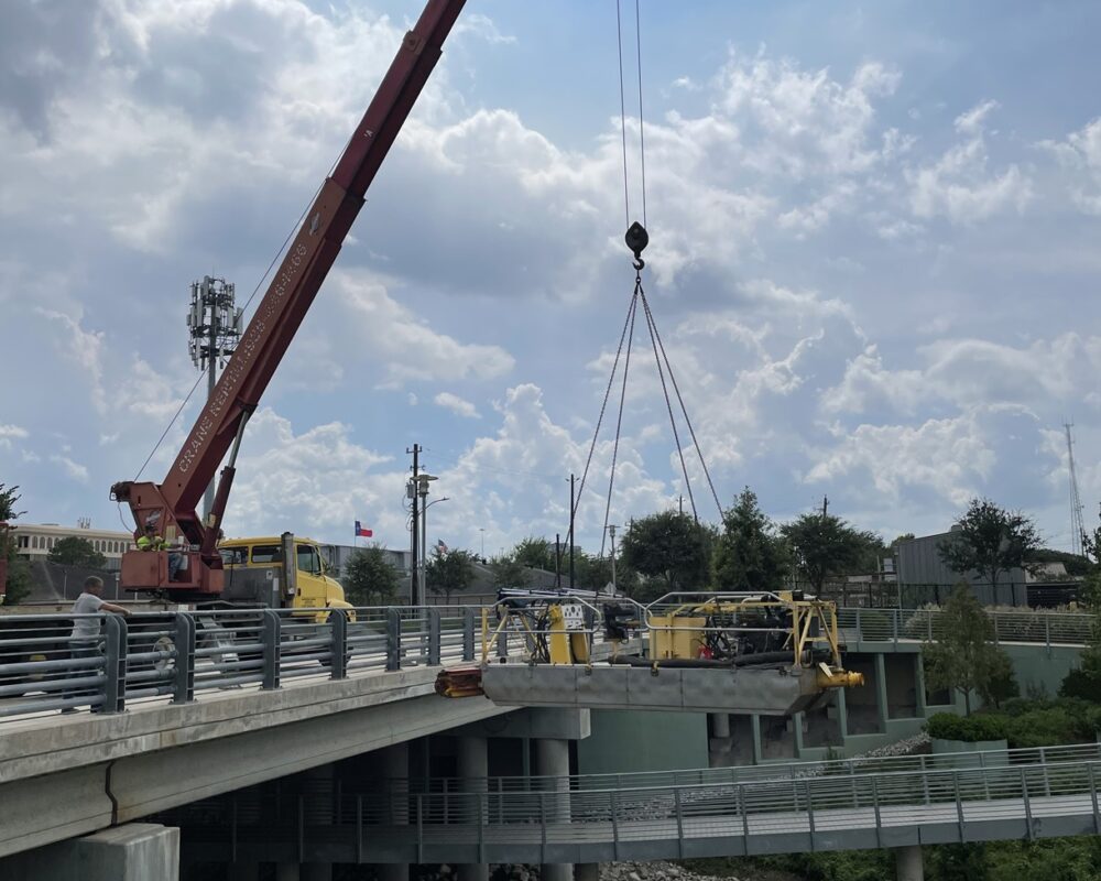 A coordinated lift of hydraulic dredging equipment being lowered into a Texas waterway using a large crane mounted on a bridge for a municipal project to remove sediment and improve water flow. The barge is a compact, specialized dredge used to fit into small spaces.